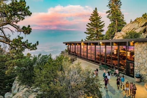 A scenic view of a mountain lodge with large windows, surrounded by trees and visitors enjoying the landscape at sunset.