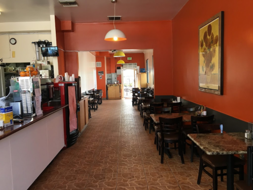 Interior of a cozy restaurant with orange walls, wooden tables, and a counter with drinks and kitchen access.