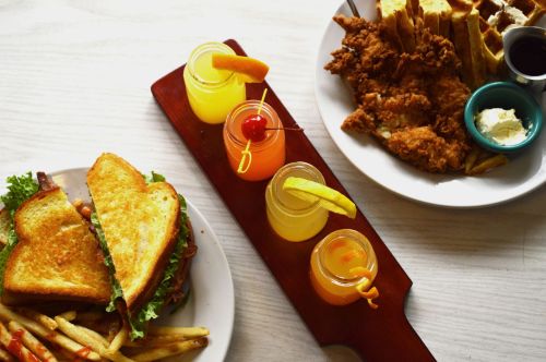 A plate with a sandwich and fries, alongside a platter of chicken and waffles with colorful drinks in shot glasses.