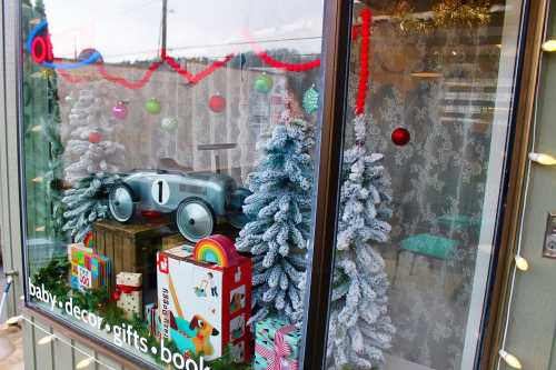 A festive shop window displays Christmas decorations, toy cars, and colorful gifts among snowy trees.