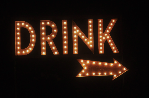 Brightly lit sign reading "DRINK" with an arrow pointing to the right, set against a dark background.