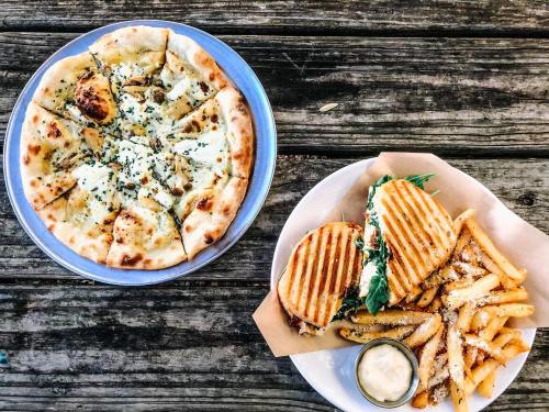 A wooden table with a pizza topped with herbs and a plate of sandwiches with fries and dipping sauce.
