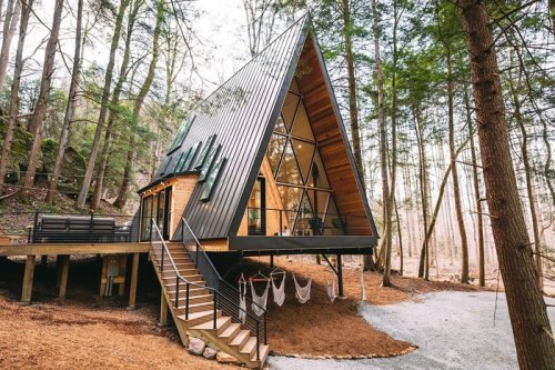 A modern A-frame cabin nestled in a forest, featuring large windows and a deck with hammocks.