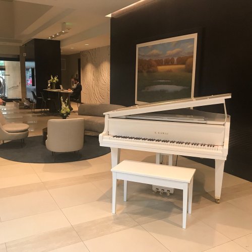 A white grand piano in a modern lobby with seating and a landscape painting on the wall.