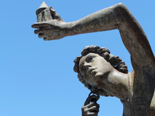 A bronze statue of a figure holding a geometric shape, set against a clear blue sky.