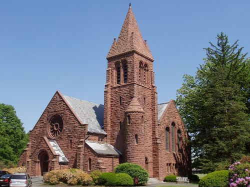 A red stone church with a tall spire, surrounded by greenery and colorful flowers under a clear blue sky.