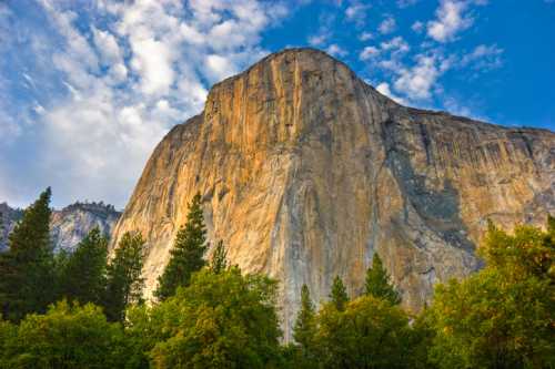 A towering rock face rises above lush green trees under a blue sky with scattered clouds.