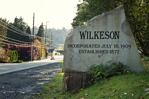 A large stone sign reading "Wilkeson" with incorporation and establishment dates, set along a quiet road.