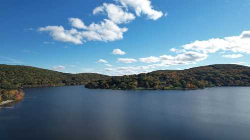 A serene lake surrounded by rolling hills and colorful autumn foliage under a clear blue sky with fluffy clouds.