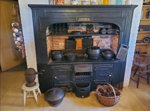 A vintage black kitchen stove with pots, a basket, and various utensils displayed in a rustic setting.