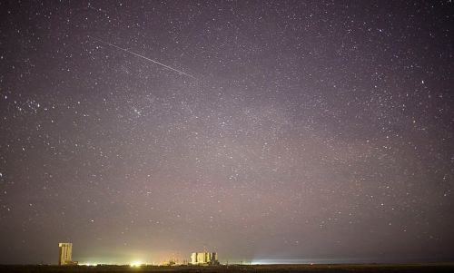 A starry night sky with a faint meteor streaking above a distant landscape and illuminated structures.