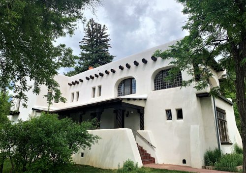 A white adobe-style building with arched windows, surrounded by greenery and under a cloudy sky.