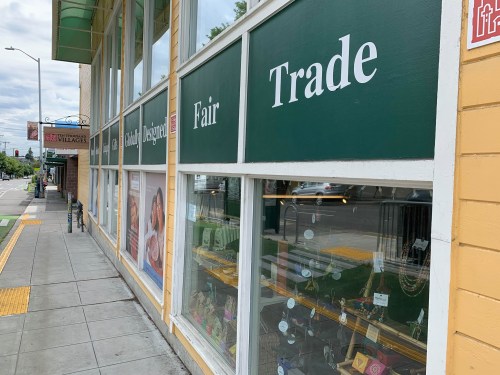 Storefront with green signs reading "Fair Trade" and "Designer" along a sidewalk. Display of items visible in windows.