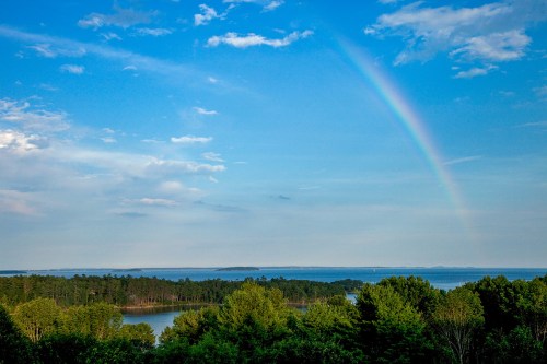 A vibrant rainbow arcs over a serene landscape of trees and a calm lake under a blue sky with scattered clouds.