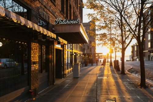 Sunset casts warm light on a brick building with a sign, as people stroll along a tree-lined street.