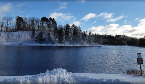 A serene winter landscape featuring a snowy shoreline and a calm lake surrounded by trees under a blue sky.