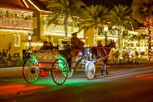 A horse-drawn carriage decorated with lights rides past festive buildings at night, surrounded by palm trees.