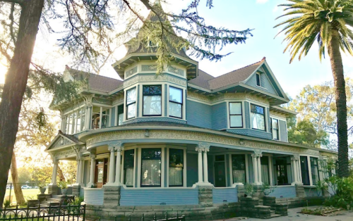 A large, ornate Victorian-style house with a blue exterior, multiple windows, and palm trees in the background.