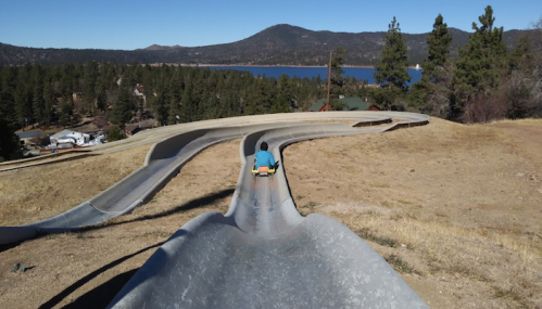 A person slides down a winding track on a sunny day, with mountains and a lake in the background.