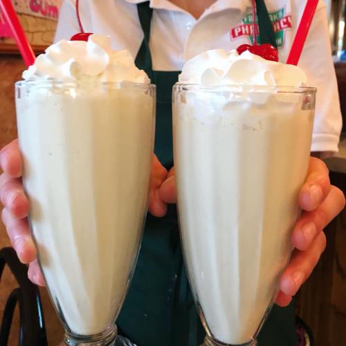 Two tall glasses of milkshakes topped with whipped cream and cherries, held by a person in an apron.