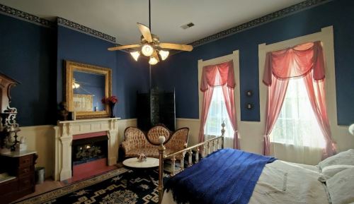 Cozy bedroom with blue walls, vintage furniture, a fireplace, and large windows with pink drapes.