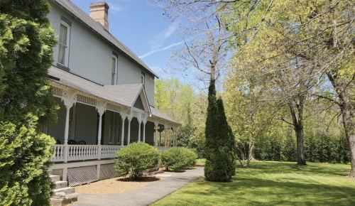 A historic house with a porch, surrounded by lush greenery and trees under a clear blue sky.