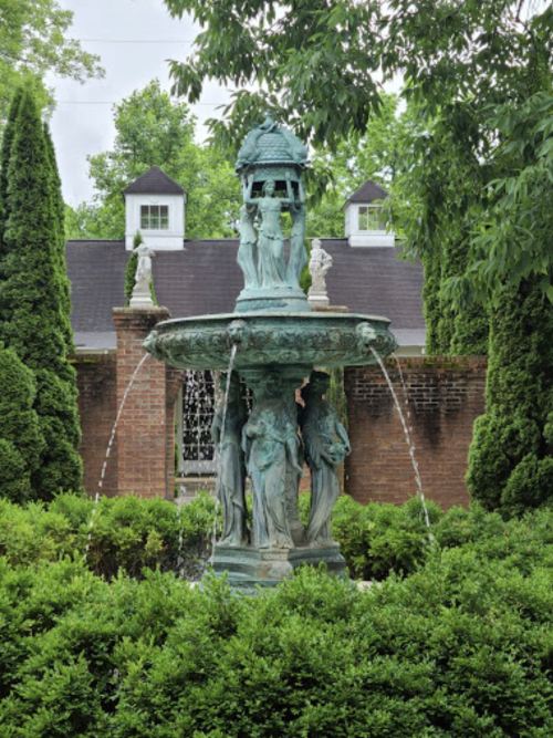 A decorative fountain surrounded by lush greenery and trees, with water flowing from its ornate sculptures.