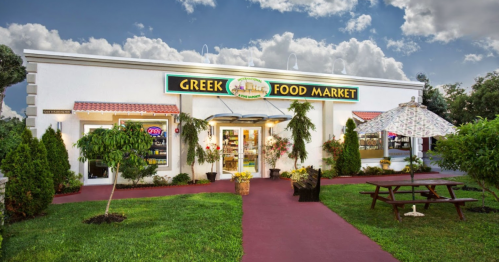 Exterior of a Greek food market with a colorful sign, surrounded by greenery and a picnic table in front.