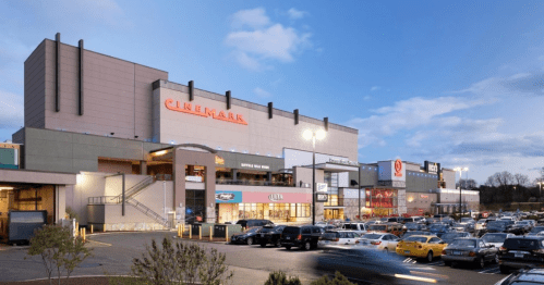 A Cinemark theater building with shops and a busy parking lot under a blue sky at dusk.