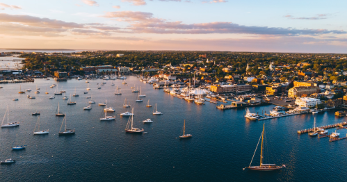 Aerial view of a coastal town with numerous boats in the harbor and a sunset sky, showcasing vibrant buildings along the shore.