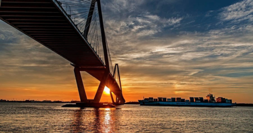 A bridge spans over water at sunset, with a cargo ship passing beneath and vibrant clouds in the sky.
