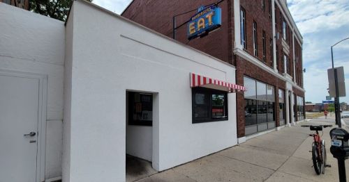 A small white building with a red-striped awning and a sign that says "EAT," next to a brick building.