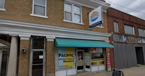 Exterior of Pacific International Market, featuring a green awning and an "Open" sign, with brick buildings nearby.