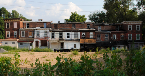 Abandoned brick building with boarded windows, overgrown vegetation, and a clear blue sky in the background.