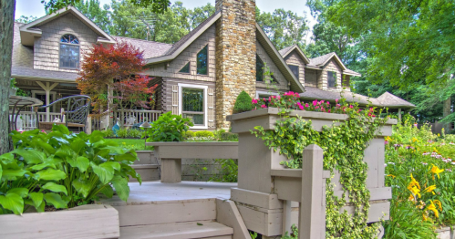 A charming house with a stone chimney, surrounded by lush greenery and colorful flowers in a vibrant garden.