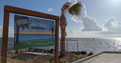 Sign for Cypremort Point State Park in Louisiana, with a view of the water and palm trees in the background.