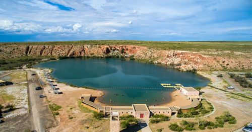Aerial view of a serene lake surrounded by cliffs, with a sandy area and buildings near the water's edge.
