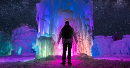 A person stands before colorful ice formations illuminated under a starry night sky.