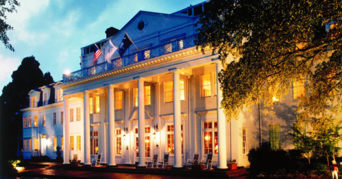 A grand white mansion with columns, illuminated windows, and rocking chairs on the porch, set against a twilight sky.