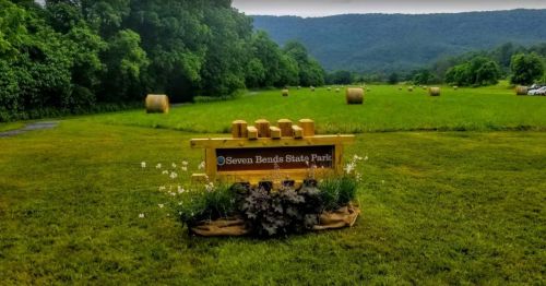 Sign for Seven Bends State Park in a grassy field with hay bales and mountains in the background.