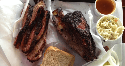 A plate of smoked beef ribs with a side of coleslaw, barbecue sauce, and slices of bread on parchment paper.