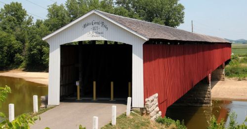 A red and white covered bridge over a calm river, surrounded by greenery and fields under a clear blue sky.