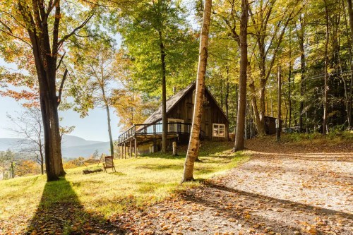 A cozy cabin surrounded by colorful autumn trees and a winding path, with a chair on the lawn.