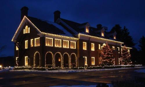 A beautifully lit brick house adorned with Christmas lights and decorated trees, set against a dark evening sky.