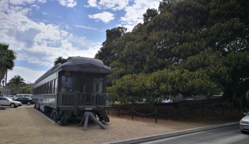 A vintage train car parked beside a large tree, with cars and a blue sky in the background.