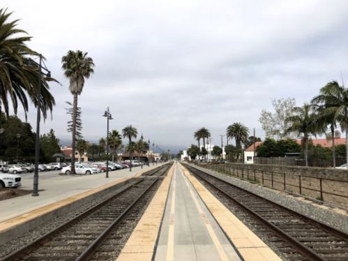 View of a train track lined with palm trees, leading towards a cloudy sky and distant mountains.