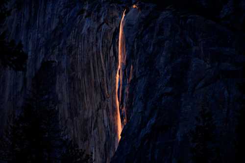 A glowing waterfall cascades down a dark rock face, illuminated by the setting sun.