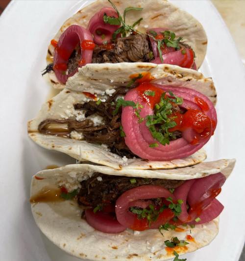 Three tacos filled with shredded meat, topped with pickled onions, cilantro, and drizzled with sauce on a white plate.