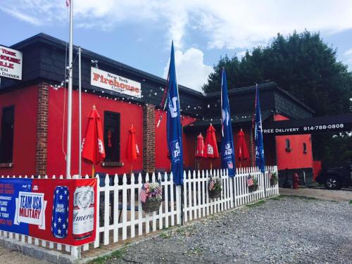 A red building with flags and umbrellas, featuring a white picket fence and signs for free delivery and military support.