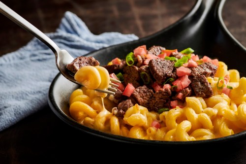 A fork lifts macaroni and cheese topped with diced steak and tomatoes from a black skillet, with a blue cloth in the background.